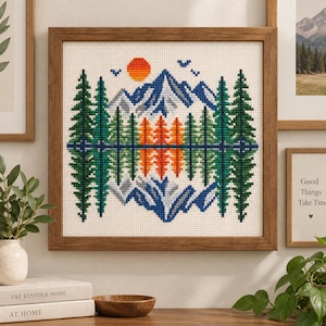 Könnte beinhalten: Ein quadratisches Kreuzstich-Stickbild in einem Holzrahmen, das eine symmetrische Berglandschaft mit grünen Tannen, blauen Berggipfeln, einer orangefarbenen Sonne und Vögeln zeigt, die sich an einer horizontalen Linie auf weißem Stoff spiegeln.