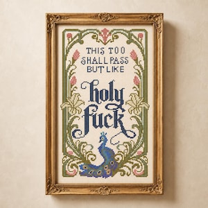 May include: A framed cross-stitch embroidery featuring the text "THIS TOO SHALL PASS BUT LIKE holy fuck" in dark blue lettering. The design is surrounded by a decorative border of green vines, pink flower buds, and a blue peacock at the bottom.