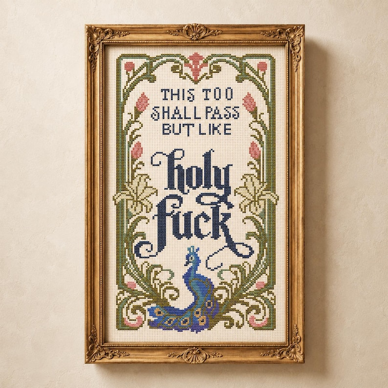 May include: A framed cross-stitch embroidery featuring the text "THIS TOO SHALL PASS BUT LIKE holy fuck" in dark blue lettering. The design is surrounded by a decorative border of green vines, pink flower buds, and a blue peacock at the bottom.