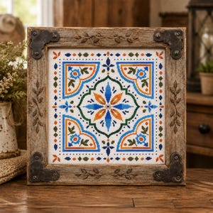 Peut inclure: Une broderie au point de croix carrée présentant un motif géométrique symétrique en fils bleus, orange et verts sur tissu blanc, encadrée dans un cadre en bois vieilli avec des coins en métal foncé et des détails de feuilles en relief.