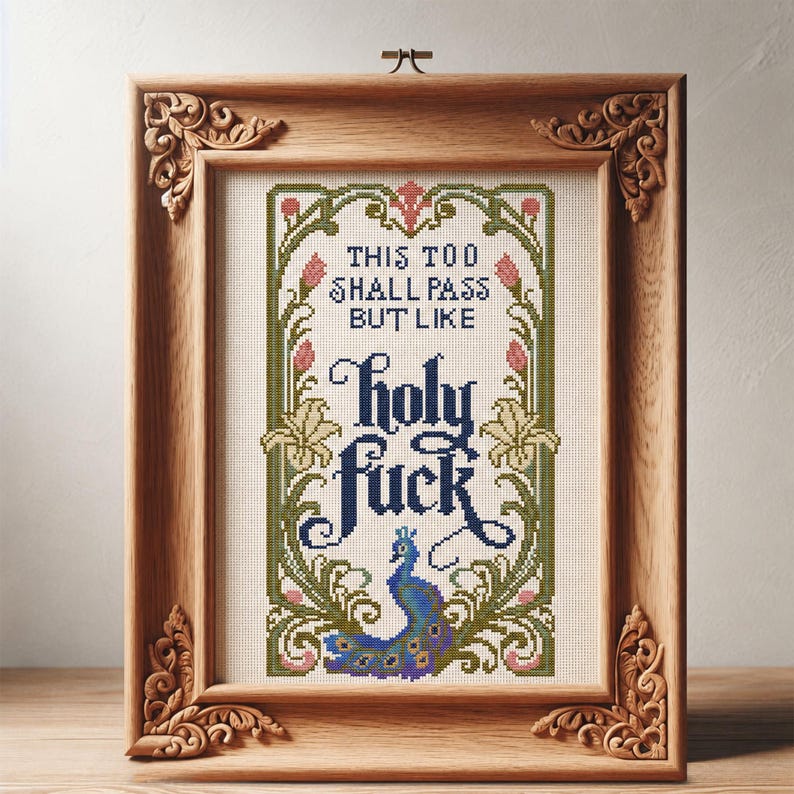 May include: A cross-stitch embroidery in a light brown wooden frame with ornate corner carvings. The design features a peacock at the bottom, floral borders in green and pink, and the text: THIS TOO SHALL PASS BUT LIKE holy fuck.