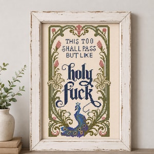 May include: A framed cross-stitch embroidery on cream fabric featuring a decorative green and pink floral border with a peacock at the bottom. The text reads: THIS TOO SHALL PASS BUT LIKE holy fuck, rendered in black and dark blue lettering.