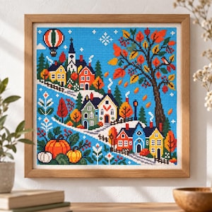 Könnte beinhalten: Eine gerahmte Kreuzstichstickerei, die eine Dorfszene mit bunten Häusern, einem großen Baum mit orangefarbenen und roten Herbstblättern, Kürbissen und einem Heißluftballon vor einem hellblauen Himmel zeigt, in einem hellen Holzrahmen.