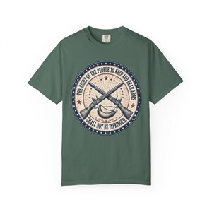 May include: A sage green short-sleeve t-shirt featuring a circular graphic with two crossed muskets and a powder horn. The text reads, "THE RIGHT OF THE PEOPLE TO KEEP AND BEAR ARMS SHALL NOT BE INFRINGED," surrounded by a border of blue stars.