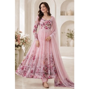 May include: A floor-length light pink Anarkali dress featuring a scoop neckline and long sleeves. The bodice and skirt are adorned with a vibrant floral print in shades of pink, purple, and green. A matching sheer pink dupatta is draped over one shoulder.