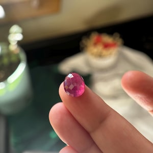 May include: A small, oval-shaped, faceted gemstone with a deep magenta-pink color. The stone features a polished surface with multiple geometric facets that reflect light, creating a sparkling appearance against the skin of a finger.