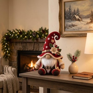 May include: A crocheted gnome figurine featuring a tall, pointed red hat decorated with white snowflake patterns, a beige flower, green leaves, and red berries. The gnome has a long white beard, a beige nose, and a red and beige crocheted outfit.