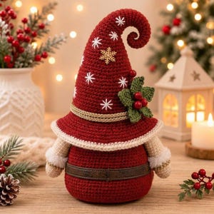 May include: A crochet gnome figurine featuring a tall, pointed red hat adorned with white embroidered snowflakes and a gold metallic trim. The gnome has a dark red body, a brown belt, and green holly leaves with red berries attached to the hat.
