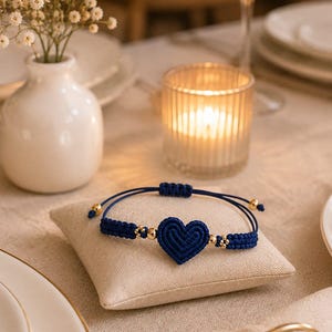 May include: A navy blue cord bracelet featuring a central heart-shaped woven knot, accented with small gold-toned beads on either side. The bracelet includes an adjustable sliding knot closure made from matching navy blue cord for a secure fit.