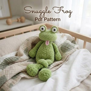 May include: A light green crocheted frog toy with large white eyes and a small pink tongue. The text Snuggle Frog Pdf Pattern is visible above the frog. The frog has a textured yarn surface and is positioned with its arms and legs crossed.