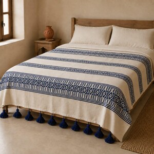 Peut inclure: Un couvre-lit couleur crème orné de rayures horizontales bleu marine aux motifs géométriques complexes. Le bord inférieur du textile est agrémenté d'une rangée de pompons bleu marine suspendus, recouvrant un cadre de lit en bois avec deux oreillers.