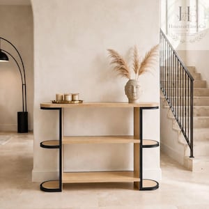 May include: A light wood console table with three tiers and black metal side supports. On the top shelf, there is a gold tray holding three candles and a beige face-shaped vase containing dried pampas grass stems.