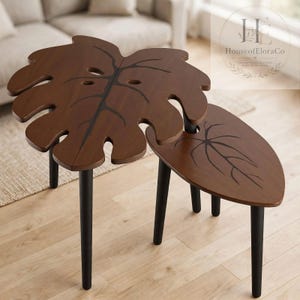 May include: A set of two nesting tables with dark brown wooden tops shaped like monstera and teardrop leaves, featuring black painted veins and sturdy black cylindrical legs. The larger table has a split leaf design with cutouts.