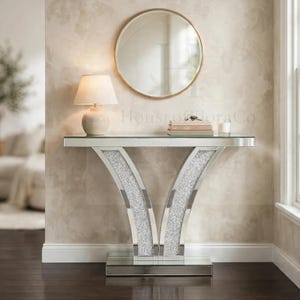 May include: A mirrored console table featuring two curved, V-shaped legs filled with crushed silver crystals. The table has a rectangular glass top and a mirrored base. A round gold-rimmed mirror hangs above the table, which holds a lamp.