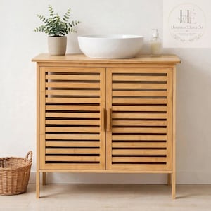 May include: A light brown bamboo bathroom vanity cabinet with two slatted doors and vertical handles. A white ceramic vessel sink sits on top, accompanied by a small potted green plant and a clear soap dispenser. A woven basket sits nearby.