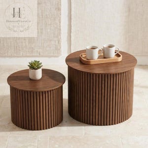 May include: Two cylindrical side tables made of dark brown wood with vertical slatted detailing. The smaller table holds a small green succulent in a white pot, while the larger table features a wooden tray with two white ceramic mugs.