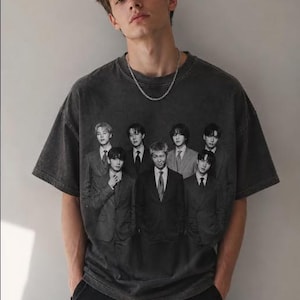 May include: A charcoal gray short-sleeve t-shirt featuring a black and white graphic print of seven individuals wearing suits and ties. The shirt has a crew neckline and a relaxed fit, paired with a simple silver chain necklace worn around the neck.