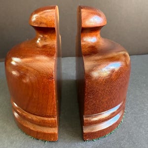May include: A pair of polished, reddish-brown wooden bookends featuring a smooth, rounded top section and a wider, curved base. Each piece has a flat vertical side and a distinct grain pattern, with a green felt lining visible along the bottom edge.