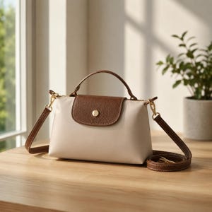 May include: A beige handbag featuring a brown pebbled leather flap with a gold-tone snap closure, a top handle, and a detachable brown leather shoulder strap with gold-tone hardware clips, presented in a structured, compact design.
