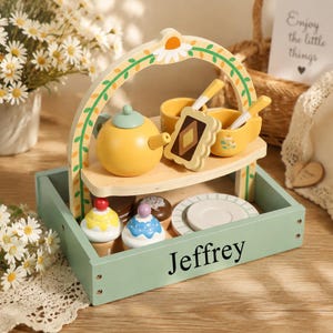 May include: A wooden toy tea set in a sage green tray with the name Jeffrey printed on the side. The set includes a yellow teapot, two yellow cups with spoons, a biscuit, two cupcakes, and a plate, all arranged on a two-tiered stand with a floral arch.