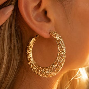 Puede incluir: Un pendiente de aro grande en color dorado que presenta un intrincado patrón de filigrana calada en toda la banda circular. La superficie metálica tiene un acabado pulido que refleja la luz, creando una apariencia texturizada en el aro.