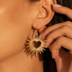 Puede incluir: Un pendiente colgante de tono dorado que presenta un recorte central en forma de corazón rodeado por un diseño de rayos de sol con numerosas puntas que se extienden hacia afuera. El pendiente cuelga de un gancho metálico simple.