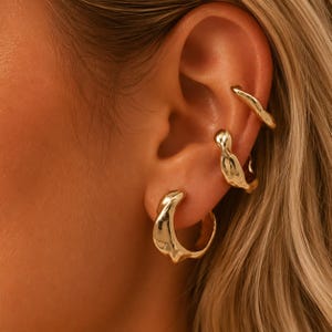 Könnte beinhalten: Ein Set aus drei goldfarbenen Ohrschmuckstücken mit unregelmäßiger, geschmolzen wirkender Textur. Das Set besteht aus einem kleinen Creolen-Ohrring am Ohrläppchen und zwei Ohrklemmen, die am oberen Knorpelbereich des Ohrs platziert sind.