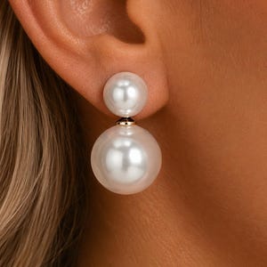 Puede incluir: Un pendiente de doble perla que presenta una perla esférica blanca más pequeña en la parte superior y una perla esférica blanca más grande suspendida debajo. Ambas perlas están conectadas por un pequeño componente metálico.