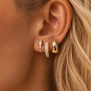 May include: Three gold-toned hoop earrings worn in a single ear. The first earring is a thin hoop with a single rectangular stone, the second is a thicker hoop encrusted with small clear stones, and the third is a smooth, polished gold dome hoop.