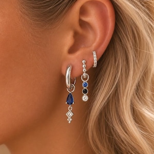 Può includere: Un orecchino a cerchio in argento con una pietra blu a goccia e un ciondolo a rombo, un orecchino pendente in argento con tre piccole pietre circolari blu, nere e trasparenti, e una clip per orecchio in argento con una fila di pietre trasparenti.