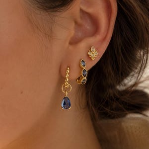 Può includere: Tre orecchini color oro indossati su un solo orecchio. L'orecchino superiore è un piccolo perno decorativo. L'orecchino centrale è un piccolo cerchio con tre pietre blu. L'orecchino inferiore è un cerchio con un ciondolo blu a forma di goccia.