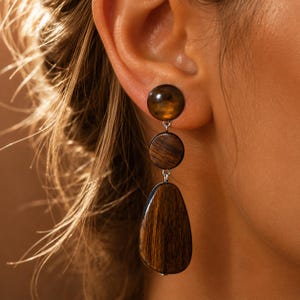 May include: A drop earring featuring three distinct components: a smooth, dark brown circular stud, a smaller dark brown circular wooden piece, and a larger, elongated, teardrop-shaped wooden pendant with a visible dark wood grain texture.