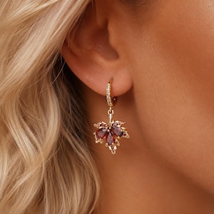 May include: A gold hoop earring featuring a dangling maple leaf charm. The leaf is adorned with deep red, pear-shaped gemstones and small clear crystals along the hoop and the leaf edges, creating a detailed and reflective jewellery piece.