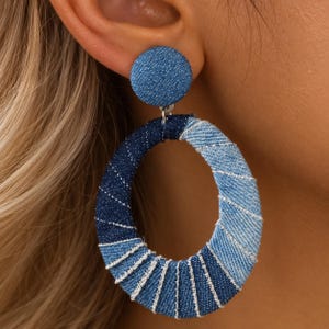 Peut inclure: Une boucle d'oreille créole circulaire présentant un design en tissu denim enroulé dans des tons alternés de bleu foncé et bleu clair. La boucle comprend un clou assorti en denim, avec des coutures blanches visibles sur le tissu.