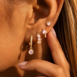 May include: A set of gold-tone earrings featuring a small heart-shaped stud with a pink stone, a small hoop with a pink heart-shaped stone, and a larger hoop with a row of small clear stones and a single round pink stone dangling from the bottom.