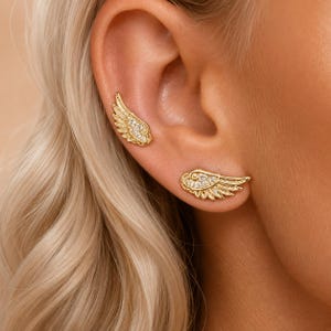May include: Two gold-toned wing-shaped earrings featuring small, clear, sparkling stones embedded along the upper edge of each wing. The earrings are positioned on the earlobe and the upper cartilage area, showcasing a detailed feather design.