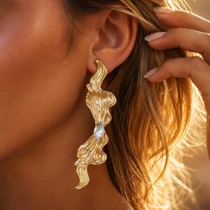 May include: A long, gold-toned earring with a wavy, organic, rippled texture. The earring features a single, clear, marquise-cut gemstone set in the centre of the elongated, flowing metal design, which hangs vertically along the side of the face.