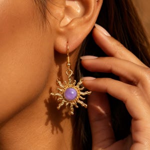 Puede incluir: Un pendiente colgante de tono dorado con un dije en forma de sol con rayos ondulados que irradian desde una piedra central redonda, lisa y de color morado claro. El pendiente cuelga de un gancho sencillo dorado con acabado metálico.