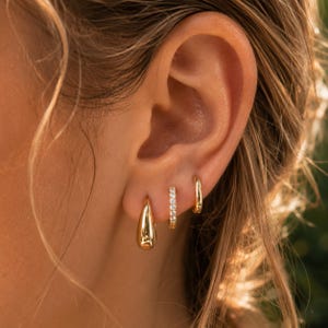 Puede incluir: Tres pendientes dorados colocados en fila en el lóbulo de la oreja. El primero es un aro liso y pulido en forma de gota. El segundo es un aro fino y estrecho incrustado con pequeñas piedras claras y brillantes. El tercero es un aro dorado sencillo y pulido.