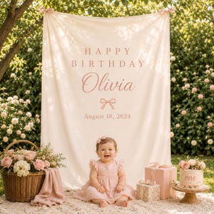 May include: A cream-colored fabric backdrop featuring the text HAPPY BIRTHDAY Olivia and August 18, 2024 in pink lettering, accented with a small pink bow graphic. The banner is suspended by two small bows at the top corners for a celebration.