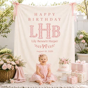 May include: A cream-colored fabric backdrop featuring the text HAPPY BIRTHDAY, the monogram LHB in a pink gingham pattern, the name Lily Bennett Harper, a bow graphic, and the date August 18, 2026, suspended by pink ribbons at the top corners.