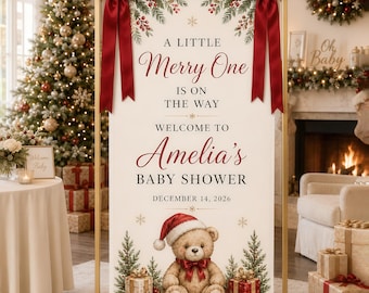 May include: A vertical rectangular sign in a gold metal frame with two large red satin bows at the top. The sign features the text A LITTLE Merry One IS ON THE WAY WELCOME TO Amelia's BABY SHOWER DECEMBER 14, 2026, a teddy bear in a Santa hat, and pine trees.