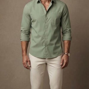 May include: A sage green long-sleeved button-down shirt with a collar and a single chest pocket, paired with cream-colored trousers. The shirt features a front button placket and is worn with the sleeves rolled up to the mid-forearm.