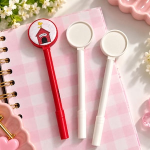 May include: Three pens with circular toppers. One pen is red and features a graphic of Woodstock sitting on a red doghouse with the text SNOOPY. The other two pens are solid white with blank circular toppers. The pens have a smooth, cylindrical design.
