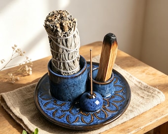 May include: A blue ceramic incense and smudge holder set featuring a large cylindrical cup for a sage bundle, a smaller cup for a palo santo stick, and a central dome-shaped incense stick holder, all decorated with a dark blue floral pattern.