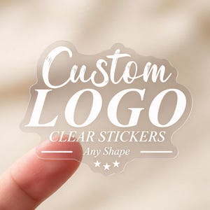 Puede incluir: Una pegatina transparente con texto blanco que dice Custom LOGO CLEAR STICKERS Any Shape, con un borde decorativo y tres pequeñas estrellas en la parte inferior. La pegatina tiene una forma troquelada y es transparente, dejando ver la superficie.