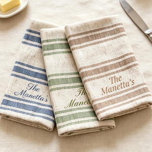 May include: Three folded fabric napkins with a striped pattern in blue, green, and brown. Each napkin features the embroidered text "The Manetta's" in a matching colour. The napkins are made of a textured, light-coloured woven material.