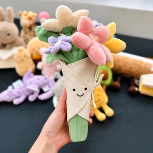 Peut inclure: Un bouquet de fleurs en peluche dans des tons rose, jaune, crème et violet, enveloppé dans un cône en tissu beige avec un visage souriant brodé. Les fleurs présentent des pétales doux et des feuilles vertes, maintenues par un ruban en tissu.