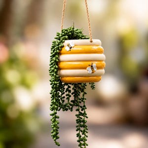 May include: A hanging ceramic planter shaped like a beehive with alternating horizontal bands of yellow and cream. Two small, raised bee figures are attached to the exterior. A green string of pearls plant cascades from the top, suspended by a rope.