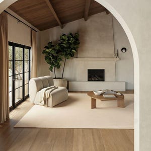 May include: A cream-colored upholstered armchair with a curved back and a draped throw blanket, positioned next to a large potted fiddle leaf fig plant and a round wooden coffee table on a light-colored area rug in a room with a fireplace.